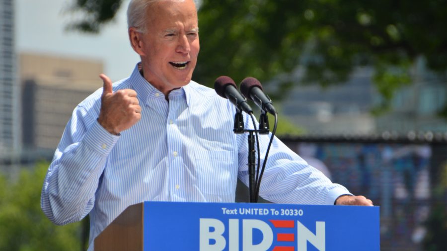 Joe Biden raises his hand in a thumbs up while giving a campaign speech at a lectern on a sunny day